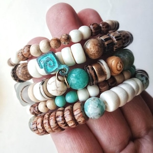 May include: A multi-strand bracelet featuring a variety of beads including smooth white, brown wood-grain, faceted turquoise, and marbled stone spheres. A small, square, oxidised teal metal charm with a spiral design hangs from the centre.