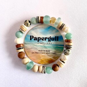 May include: A circular beaded bracelet featuring a variety of materials including light green sea glass, speckled dalmatian jasper, smooth wooden discs, and white beads. The bracelet surrounds a circular card labelled Papergull Jewellery.