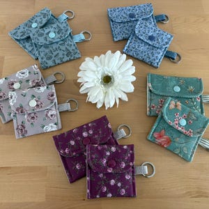 Könnte beinhalten: Eine Sammlung kleiner Stoffbeutel mit Blumenmuster und Schlüsselringen. Die Beutel sind in verschiedenen Farben wie Blau, Lila und Grün gehalten und haben Knopfverschlüsse. Eine weiße Blume befindet sich in der Mitte des Bildes.