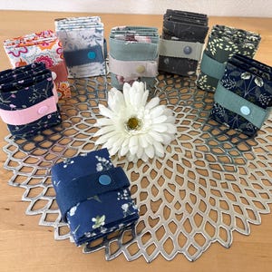 Könnte beinhalten: Eine Sammlung von acht gefalteten Stofftaschen, die jeweils mit einem passenden Gummiband und einem Kunststoff-Druckknopf verschlossen sind. Die Taschen zeigen verschiedene Blumen- und Pflanzenmuster in Farben wie Marineblau, Weiß, Grün, Grau und Rosa.