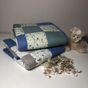 Könnte beinhalten: Eine blaue und grüne Patchwork-Decke mit Blumenmuster und einem kleinen Teddybären. Die Decke ist in der Hälfte gefaltet und hat einen Fransenbesatz.