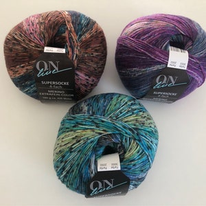 Pode incluir: Três novelos de lã em diferentes combinações de cores. Cada novelo é etiquetado com "ON live" e "SUPERSOCKE 4-fach", com o texto "MERINO EXTRAFEIN-COLOR" no novelo castanho.