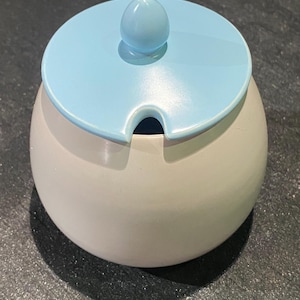 May include: A small, round sugar bowl with a pale blue lid and a matching knob. The bowl is a soft, neutral colour. The lid has a small notch for a spoon. The design is simple and elegant.