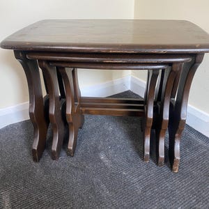 May include: A set of three dark brown wooden nesting tables with a simple design. The tables are stacked on top of each other, with the largest table on the bottom and the smallest table on top.