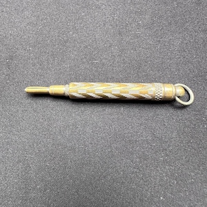 May include: A metallic pencil lead holder featuring a cylindrical body with a textured chevron pattern in gold and silver tones. The tool includes a small circular loop at one end and a split-tip mechanism at the other for holding lead.