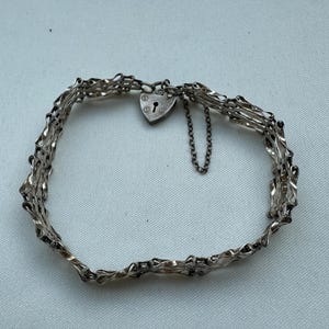 Puede incluir: Una pulsera de tono plateado con un diseño de metal tejido de múltiples hilos. La pulsera se asegura con un cierre de candado en forma de corazón que incluye una pequeña cadena de seguridad adjunta. El metal tiene un acabado pulido.