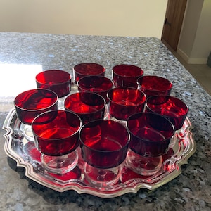 May include: A silver serving tray displays twelve ruby red glass candle holders. Each candle holder is round with a short stem. The tray has a scalloped edge and is on a granite countertop.