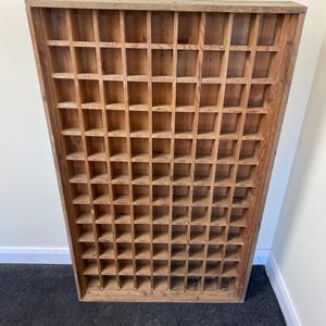 May include: A rectangular wooden wall display case featuring a grid of numerous small, square compartments. The unit is constructed from light brown wood with a visible grain pattern, designed with an open front and a solid outer frame.