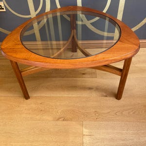 May include: A mid-century modern coffee table with a triangular wooden frame and a clear glass top. The table has a warm, natural wood finish and a unique, geometric design. The table is approximately 91 cm in diameter.