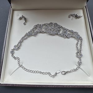 Könnte beinhalten: Ein silberfarbenes Halsketten- und Ohrringset mit filigranen Metallarbeiten und klaren Kristallakzenten. Die Halskette verfügt über ein zentrales kreisförmiges Motiv, ein doppeltes Kettendesign und einen Karabinerverschluss mit Verlängerungskette.