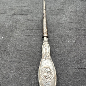 May include: A silver-toned metal button hook featuring an ornate handle with an embossed cherub face surrounded by floral detailing. The tool has a long, slender, slightly tarnished metal shaft that terminates in a small, curved hook at the top.