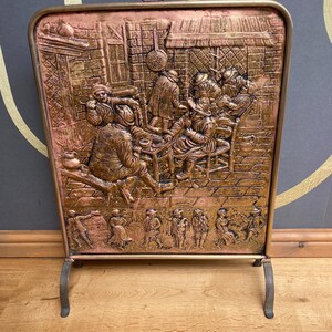 May include: A copper-toned fireplace screen featuring a detailed relief of a social gathering. The screen has a rectangular shape with a curved top and a handle. It stands on two curved legs. The screen is approximately 50 cm tall.