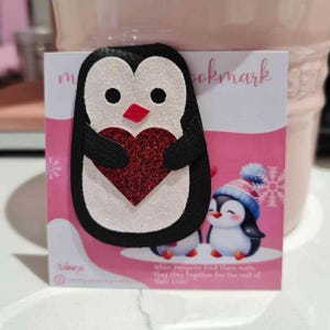 Puede incluir: Un marcapáginas de pingüino con un corazón rojo brillante. El pingüino es negro y blanco con un pico rojo. El marcapáginas tiene la palabra "bookmark" y una cita sobre pingüinos sobre un fondo rosa.