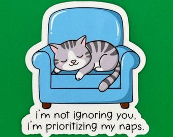 May include: A sticker featuring a cartoon cat sleeping in a blue armchair. The text reads, "I'm not ignoring you, I'm prioritising my naps."