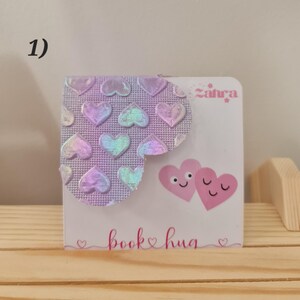Puede incluir: Un marcapáginas rosa y blanco con un diseño de corazón holográfico y el texto "book hug" en la parte inferior. El marcapáginas presenta dos corazones rosas con caras en la parte delantera.