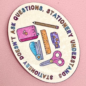 Può includere: Adesivo ovale con illustrazioni di cancelleria e la scritta "Stationery Understands, Doesn't Ask Questions". Include un taccuino rosa, una matita, un righello, nastro adesivo, una cucitrice e forbici.