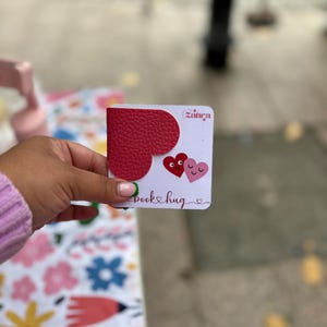 Puede incluir: Un marcapáginas de cuero rojo en forma de corazón con el texto "book hug" y un corazón rosa con una carita sonriente. El marcapáginas está hecho por Sahra.