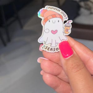 Puede incluir: Una pegatina de fantasma blanco con un corazón rosa, que lleva un gorro naranja y sostiene un café con leche de calabaza. La pegatina tiene las palabras "Pumpkin Spice Season".