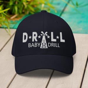 Peut inclure: Casquette de baseball bleu marine avec texte brodé blanc et un graphique de chevalet de forage. Le texte dit "D*R*L*L BABY DRILL". La casquette a une visière incurvée et une coupe structurée.