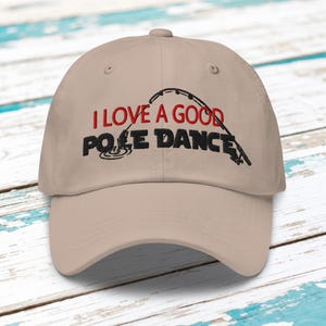 Może przedstawiać: Beżowa czapka z daszkiem z wyhaftowanym napisem I LOVE A GOOD POLE DANCE czerwoną i czarną nicią. Wzór zawiera czarny haftowany motyw wędki z żyłką, przy czym słowo POLE jest wkomponowane w grafikę przedstawiającą wędkowanie.