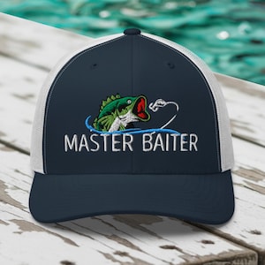 May include: Navy blue and white trucker hat with the text "MASTER BAITER" embroidered in white. The hat features a green and red fish graphic above the text, with a white fishing line design.