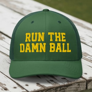May include: A green trucker cap featuring the yellow embroidered text "RUN THE DAMN BALL". The cap has a mesh back and a curved peak. The hat is displayed on a weathered wooden surface.