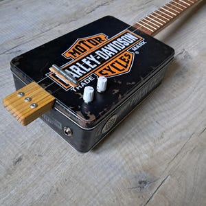 Puede incluir: Una guitarra de caja de puros con el logotipo negro de Harley-Davidson Motor Cycles en la tapa. El instrumento tiene un mástil de madera, tres cuerdas metálicas, un puente de metal, dos perillas de control blancas y un jack de entrada lateral.