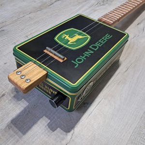 Peut inclure: Une guitare cigar box avec le logo John Deere sur un corps rectangulaire en métal noir et vert. L'instrument possède trois cordes, un manche en bois, un bouton de volume noir et un petit haut-parleur circulaire sur le panneau latéral.