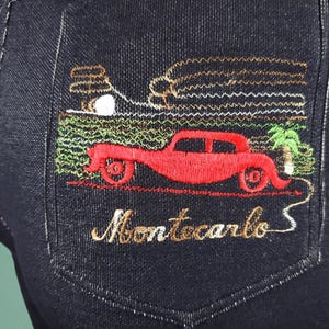 May include: Dark blue denim jeans with an embroidered pocket. The embroidery depicts a red vintage car, palm tree, and the word "Montecarlo" in gold thread. The design is detailed and colourful, adding a unique element.