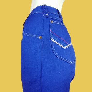 May include: Blue denim jeans with white stitching and a contrasting coloured pocket detail. The pocket has a white border and red, green, and white stitching.