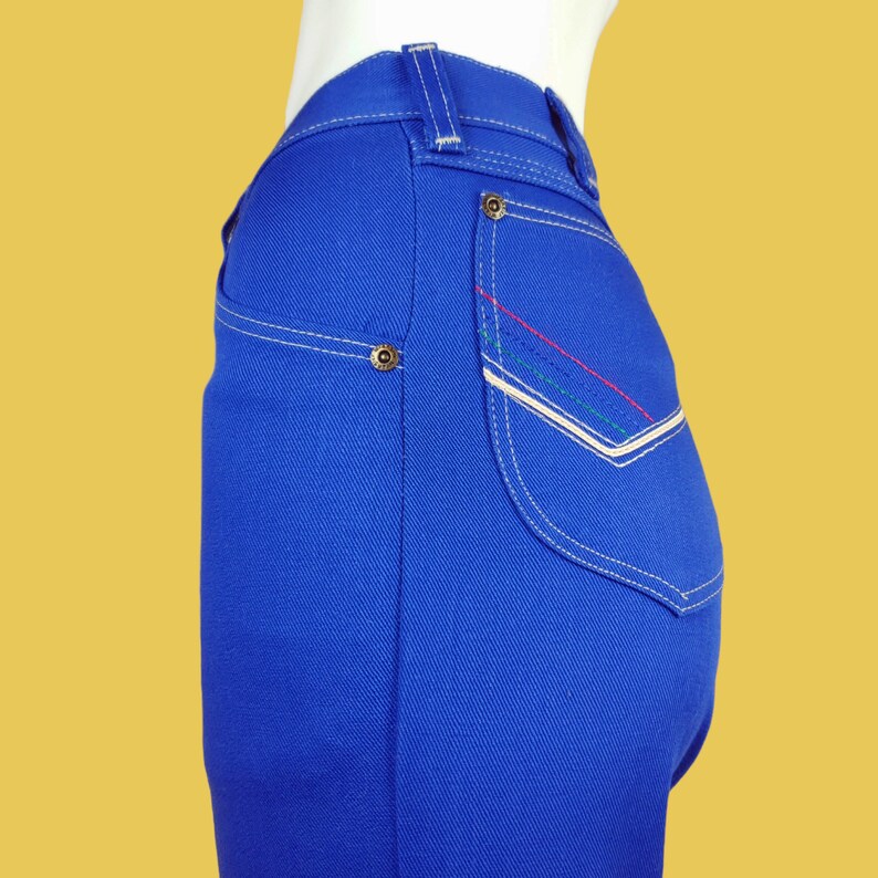 May include: Blue denim jeans with white stitching and a contrasting coloured pocket detail. The pocket has a white border and red, green, and white stitching.