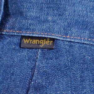 K&ouml;nnte beinhalten: Nahaufnahme von blauem Denim-Stoff mit orangefarbenen N&auml;hten. Ein schwarzes rechteckiges Etikett mit dem goldenen Wort "Wrangler" ist auf den Denim gen&auml;ht. Der Denim hat ein strukturiertes Aussehen.
