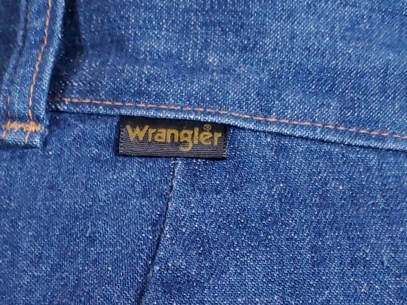 K&ouml;nnte beinhalten: Nahaufnahme von blauem Denim-Stoff mit orangefarbenen N&auml;hten. Ein schwarzes rechteckiges Etikett mit dem goldenen Wort "Wrangler" ist auf den Denim gen&auml;ht. Der Denim hat ein strukturiertes Aussehen.