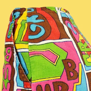 Peut inclure: Gros plan sur un pantalon &agrave; motifs color&eacute;s. Le tissu pr&eacute;sente un motif vibrant avec de grandes lettres marron, roses, vertes et bleues, sur fond blanc. Une poche est visible sur le c&ocirc;t&eacute;.