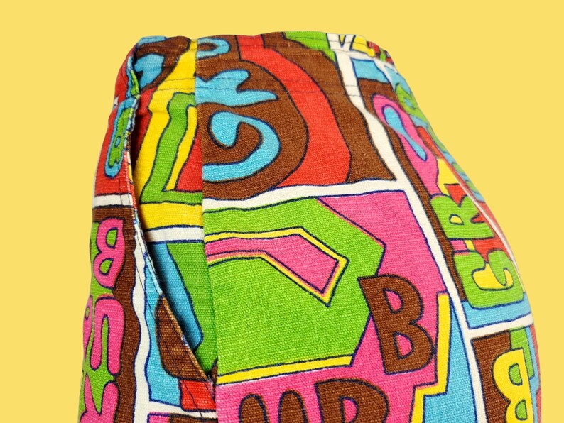 Peut inclure: Gros plan sur un pantalon &agrave; motifs color&eacute;s. Le tissu pr&eacute;sente un motif vibrant avec de grandes lettres marron, roses, vertes et bleues, sur fond blanc. Une poche est visible sur le c&ocirc;t&eacute;.