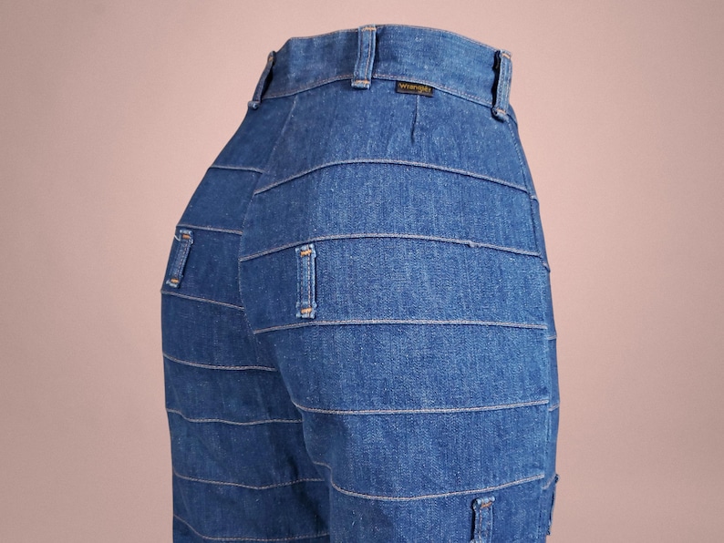 K&ouml;nnte beinhalten: Nahaufnahme einer blauen Jeans mit horizontalen Steppdetails. Die Jeans hat ein klassisches F&uuml;nf-Taschen-Design und ein Wrangler-Etikett am Bund. Die Jeans ist aus mittelblauem Denim.