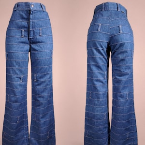 K&ouml;nnte beinhalten: Blaue Jeanshose mit hoher Taille und ausgestellten Beinen. Die Hose hat horizontale Steppdetails auf der Vorder- und R&uuml;ckseite mit kleinen rechteckigen Akzenten. Die Hose wird vor einem neutralen Hintergrund pr&auml;sentiert.