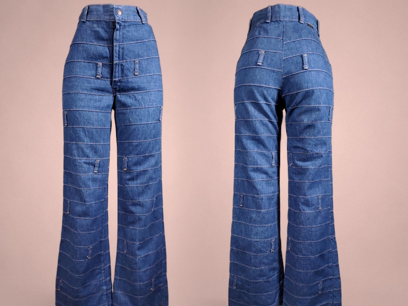 K&ouml;nnte beinhalten: Blaue Jeanshose mit hoher Taille und ausgestellten Beinen. Die Hose hat horizontale Steppdetails auf der Vorder- und R&uuml;ckseite mit kleinen rechteckigen Akzenten. Die Hose wird vor einem neutralen Hintergrund pr&auml;sentiert.