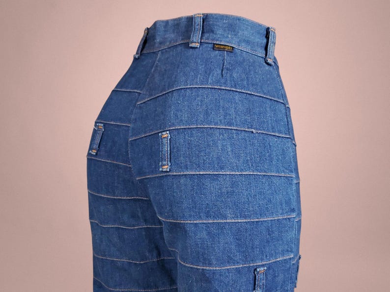 K&ouml;nnte beinhalten: Nahaufnahme einer blauen Jeans mit horizontalen Steppdetails. Die Jeans hat ein klassisches F&uuml;nf-Taschen-Design und ein Wrangler-Etikett am Bund. Die Jeans ist aus mittelblauem Denim.