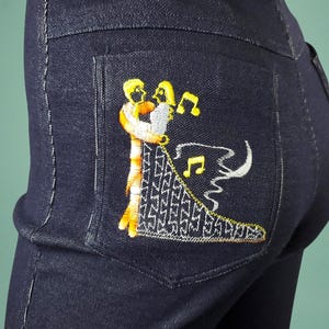 May include: Dark blue denim jeans with an embroidered design on the back pocket. The embroidery features two figures embracing, with musical notes and a decorative pattern in yellow, white, and orange.