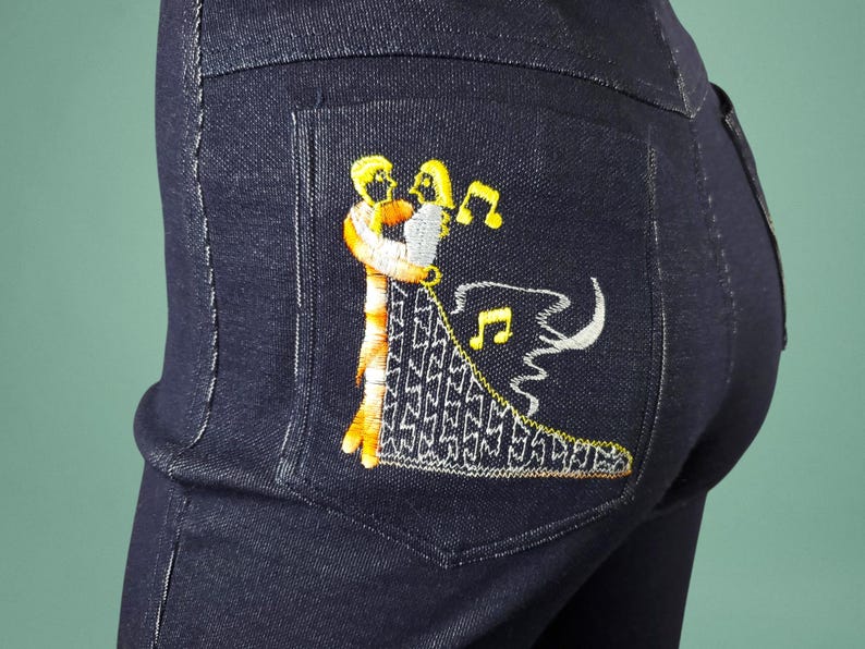 May include: Dark blue denim jeans with an embroidered design on the back pocket. The embroidery features two figures embracing, with musical notes and a decorative pattern in yellow, white, and orange.