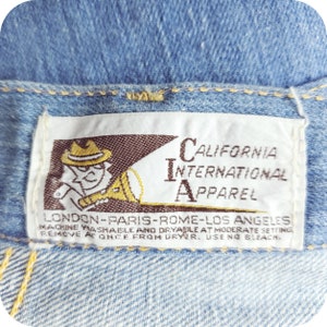 K&ouml;nnte beinhalten: Ein wei&szlig;es Etikett mit braunem Text und einer Cartoon-Illustration eines Mannes mit Hut, der ein Megaphon h&auml;lt. Der Text lautet "California International Apparel" und "London-Paris-Rom-Los Angeles". Das Etikett enth&auml;lt auch Pflegehinweise f&uuml;r Maschinenw&auml;sche und -trocknung.