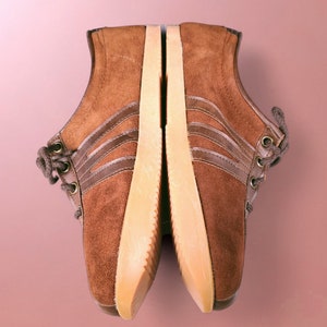 May include: A pair of brown suede sneakers with white soles and three brown stripes on each shoe.