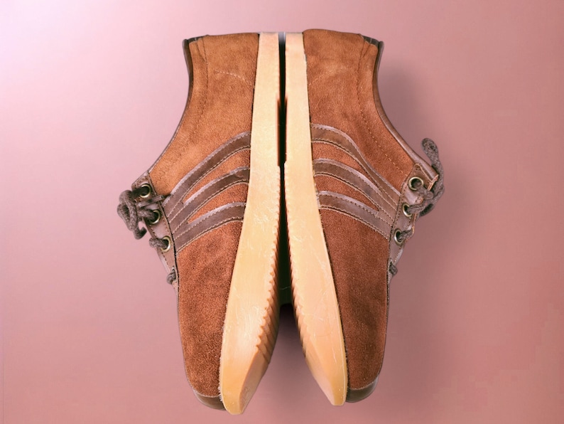 May include: A pair of brown suede sneakers with white soles and three brown stripes on each shoe.