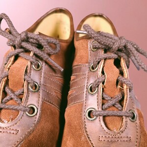 May include: A pair of brown suede shoes with brown leather accents and laces. The shoes have a classic design with a rounded toe and a lace-up closure.