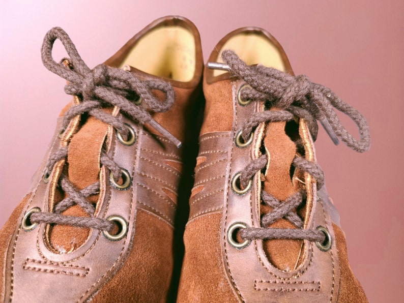 May include: A pair of brown suede shoes with brown leather accents and laces. The shoes have a classic design with a rounded toe and a lace-up closure.