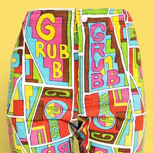 Peut inclure: Gros plan sur un pantalon color&eacute; avec un motif g&eacute;om&eacute;trique et le mot "GRUBB" en grandes lettres. Le pantalon pr&eacute;sente une fermeture boutonn&eacute;e et un m&eacute;lange de couleurs vives, notamment rose, bleu, jaune et marron.