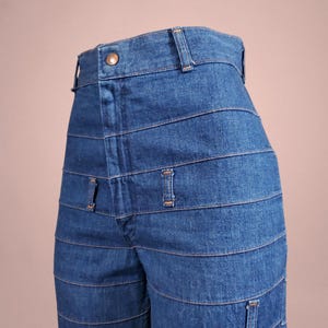 May include: Close-up of blue denim jeans with horizontal seam detailing. The jeans have a button closure and belt loops at the waist. The design features multiple horizontal seams across the legs, creating a unique, layered look.