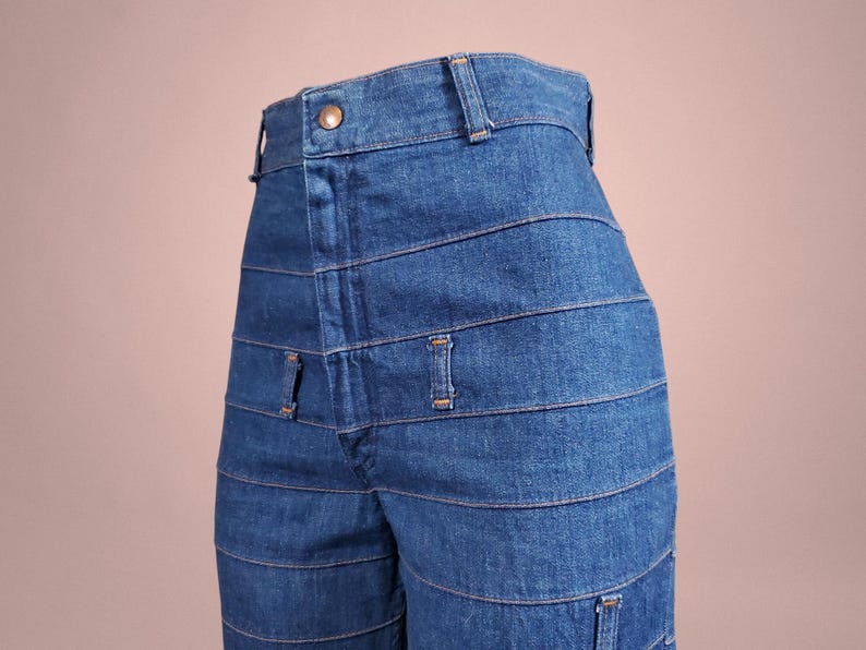 K&ouml;nnte beinhalten: Nahaufnahme einer blauen Jeans mit horizontalen Nahtdetails. Die Jeans hat einen Knopfverschluss und G&uuml;rtelschlaufen am Bund. Das Design weist mehrere horizontale N&auml;hte &uuml;ber den Beinen auf.