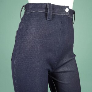 May include: Close-up of dark blue denim trousers. The trousers feature a high waist, belt loops, and a button closure. The fabric has a subtle texture, and the stitching is visible. These are a classic style, suitable for various outfits.